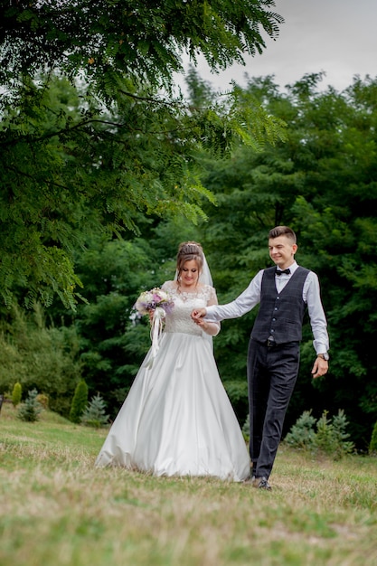 Feliz novia y el novio tomados de la mano y caminando en el día de la boda del jardín. Vista posterior de encantadores recién casados con estilo tomados de la mano mientras camina en el bosque del parque, felices momentos de matrimonio