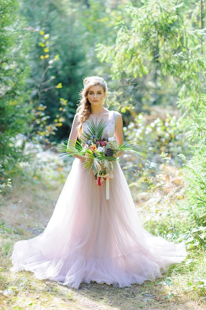 Feliz novia en un vestido de novia rosa. La niña sostiene un ramo de novia en sus manos. Ceremonia de boda de estilo boho en el bosque.