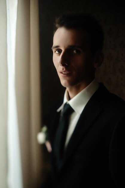 Feliz novio elegante preparándose por la mañana a la luz de la ventana en una habitación rica