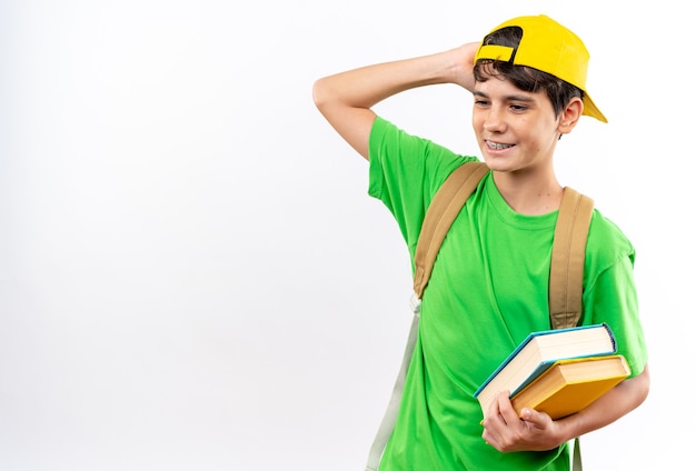Feliz, olhando para o lado, jovem estudante usando uma mochila com tampa, segurando livros isolados na parede branca com espaço de cópia