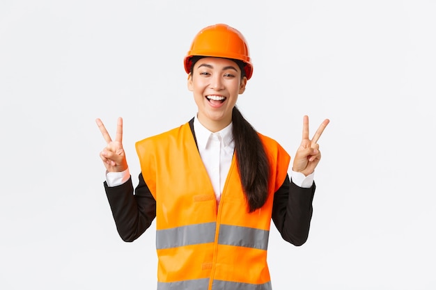 Feliz otimista arquiteta asiática feminina com capacete de segurança e jaqueta reflexiva, mostrando o símbolo da paz e sorrindo, garantida na vitória de sua empresa, vencendo o concurso em obras de construção