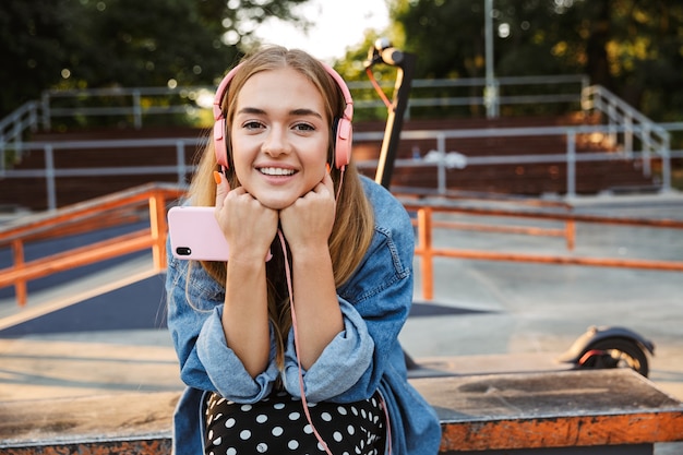 feliz otimista positiva jovem adolescente do lado de fora no parque ouvindo música com fones de ouvido segurando o telefone móvel.