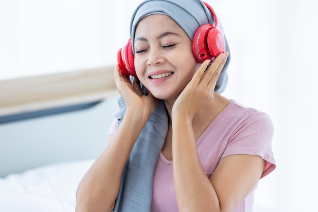 Feliz un paciente de cáncer de mama con enfermedad de las mujeres asiáticas con cinta rosa con pañuelo en la cabeza en auriculares está escuchando música después del tratamiento de quimioterapia sentarse en la cama en el dormitorio de la casa