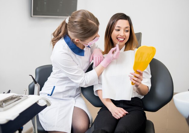 Feliz paciente y dentista con sonda están revisando los dientes en el espejo después del tratamiento dental en el consultorio dental moderno