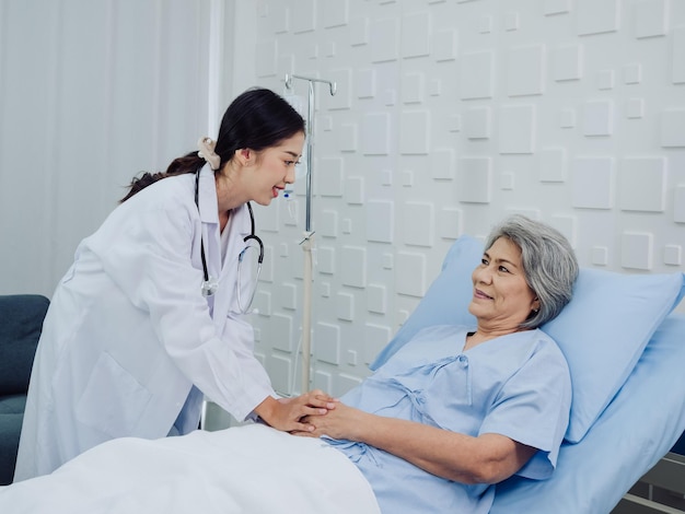 Feliz paciente idosa asiática de vestido azul claro, deitada na cama em solução salina, foi encorajada pela jovem médica de terno branco com estetoscópio e segurando a mão no quarto do hospital