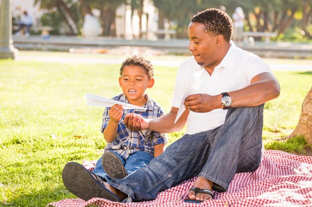 Feliz padre afroamericano y hijo de raza mixta jugando con aviones de papel en el parque