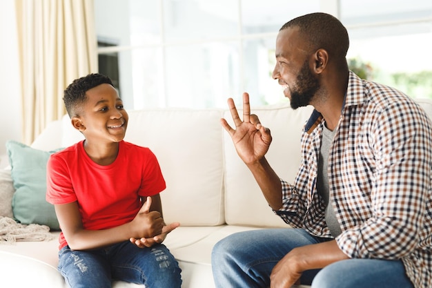 Feliz padre afroamericano con su hijo sentado en el sofá de la sala hablando en lenguaje de señas