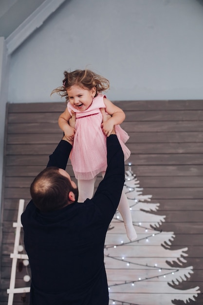 Feliz padre atractivo sostiene en sus brazos y se divierte con su pequeña casa linda hija en la atmósfera de Año Nuevo