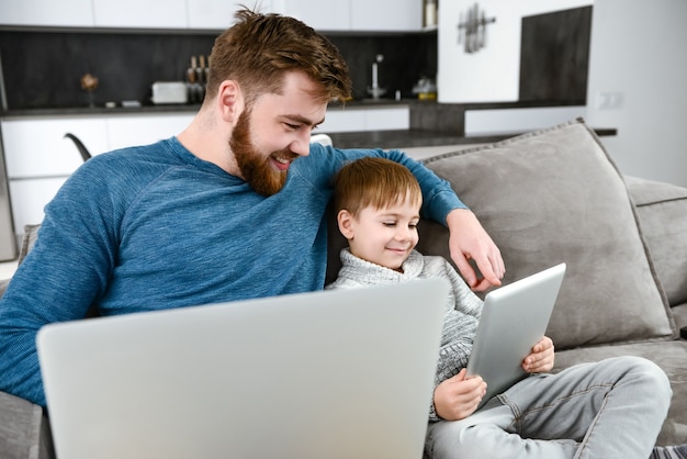 Feliz padre barbudo vestido con un suéter azul abrazando a su hijo y usando la computadora portátil mientras su hijo juega con la tableta