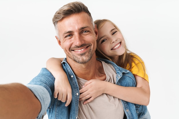 Feliz padre caucásico llevar a cuestas a su pequeña hija mientras toma selfie foto juntos aislado en blanco