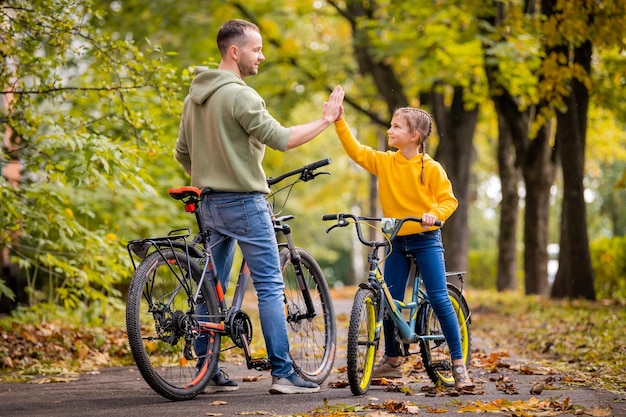 Feliz padre e hija caminan con bicicletas en el parque de otoño en un día soleado