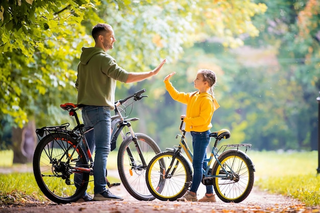 Feliz padre e hija caminan con bicicletas en el parque de otoño en un día soleado