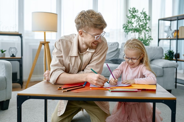 Feliz padre e hija dibujando sentados en la mesa del escritorio, papá ayudando a un niño en edad preescolar a hacer la tarea