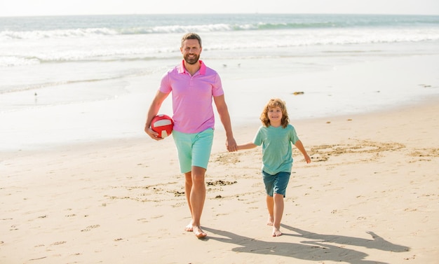 Feliz padre e hijo caminan en la playa de verano con pelota, familia.