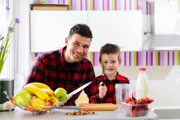 Feliz padre e hijo haciendo desayuno saludable juntos. Sentado en una cocina con cuchillo en sus manos y mirando a cámara.