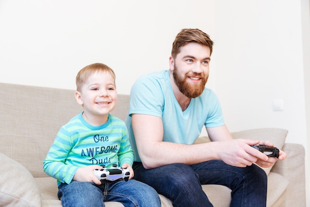 Feliz padre e hijo pequeño sentado y jugando videojuegos en el sofá en casa