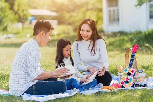 Feliz padre de familia joven asiático, madre e hijo niña divirtiéndose y disfrutando al aire libre en un libro de lectura de manta de picnic en el parque en tiempo soleado, concepto de primavera de ocio de verano