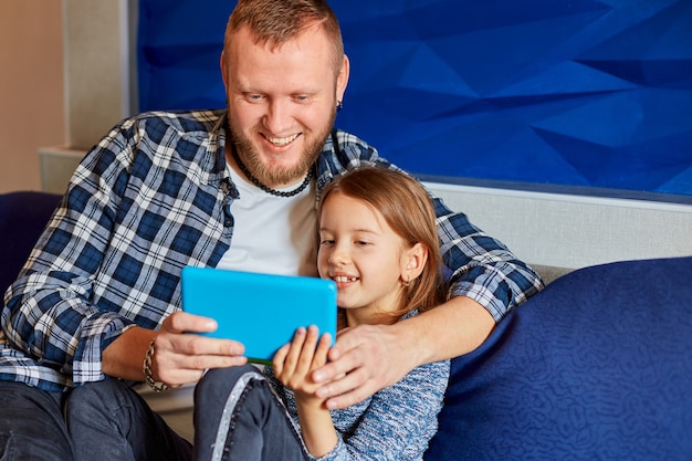 Feliz padre con hija con tablet PC en la sala de estar, en el sofá en casa, leyendo o jugando desde la tableta. Familia feliz.