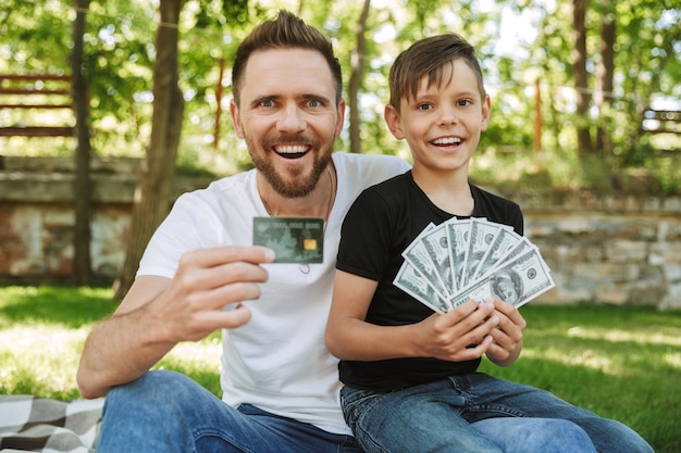 Feliz padre joven emocionado sentado con su pequeño hijo con dinero y tarjeta de crédito.