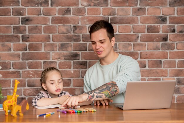 Feliz padre joven inconformista con tatuaje disfrutando de juego educativo con el niño en casa: él contando lápices de colores junto con la hija