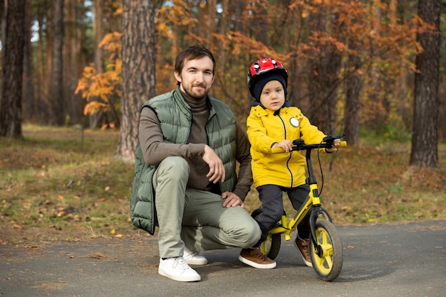 Feliz padre joven en ropa casual en cuclillas junto a su adorable hijo pequeño en casco de seguridad mientras ambos disfrutan de un cálido fin de semana en un entorno natural