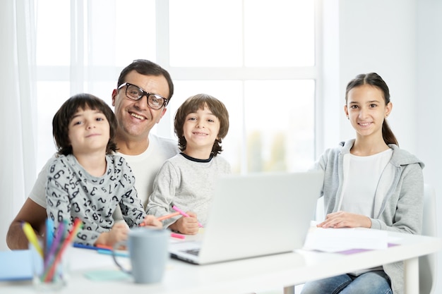 Feliz padre latino y niños sonriendo a la cámara, sentados juntos en la mesa en casa. Hombre usando laptop, trabajando desde casa y viendo a los niños. Freelance, concepto de familia