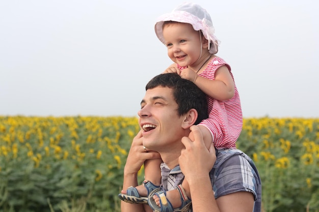 Feliz padre riendo sosteniendo a la hija bebé en los hombros de papá de pie juntos en el prado de girasoles disfrutando del verano caminando juntos en el día del padre
