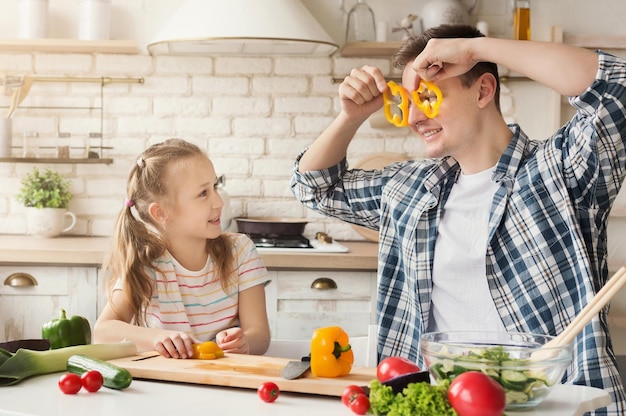 Feliz padre y su linda hija cubriendo los ojos con pimienta, divirtiéndose mientras preparan una comida saludable juntos en la cocina, espacio de copia