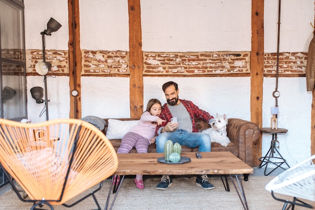 Feliz padre con su pequeña hija mirando por teléfono en casa sentado en el sofá con el perro