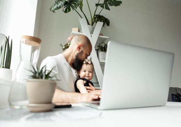 Feliz padre trabajador barbudo con una hija pequeña usa una computadora portátil mientras se sienta a la mesa en una habitación moderna