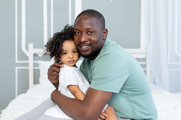 Feliz pai afro-americano abraça sua filhinha na cama em casa, família feliz