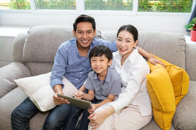 Feliz pai da família asiática mãe e filho sentados no sofá usando tablet juntos na sala de estar olhando para a câmera