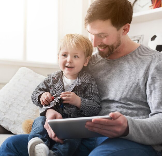 Feliz pai e filho brincando em um tablet digital, sentados no sofá em casa