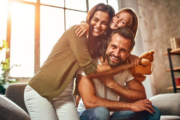 Foto feliz pai e mãe com sua filha fofa e ursinho de pelúcia abraçam e se divertem sentados no sofá da sala de estar em casa.