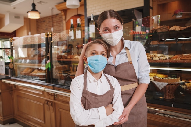 Feliz panadera y su pequeño hijo con mascarillas médicas, trabajando en la panadería de su familia durante la pandemia de coronavirus