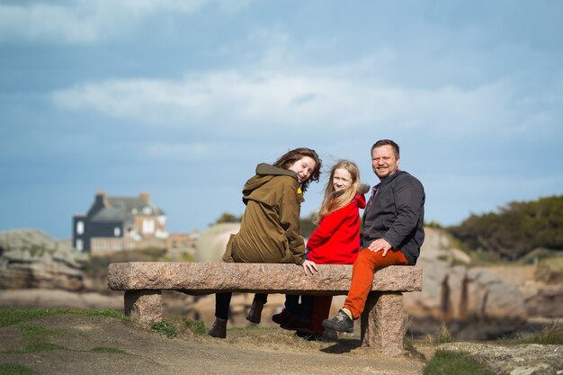 Feliz papá y dos hijas se sientan en un banco cerca del mar y la orilla en Tregastel, Bretaña. Francia