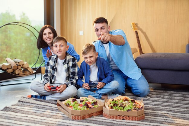 Feliz papá mamá y dos hijos sentados en el suelo jugando videojuegos con gamepads y comiendo pizza sabrosa