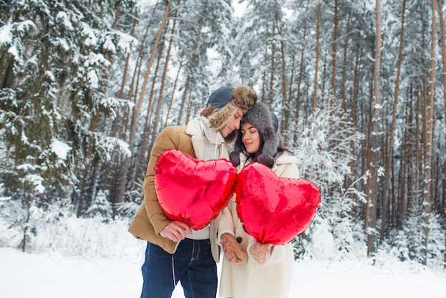 Feliz pareja abrazándose y besándose al aire libre en el bosque de invierno. Globos en forma de corazón