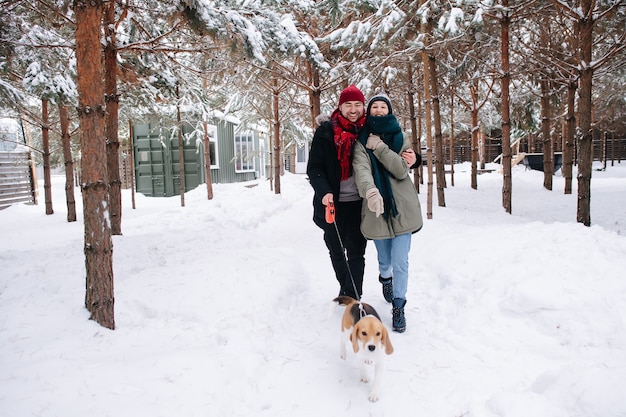 Feliz pareja abrazo caminando por un camino cubierto de nieve con su perro entre los troncos de los árboles de coníferas a través de su patio. Ambos con sombreros, chaquetas y bufandas.