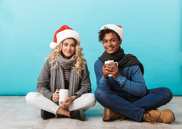 Feliz pareja de adolescentes multirraciales sentado aislado sobre la pared azul, con sombreros de navidad, sosteniendo tazas