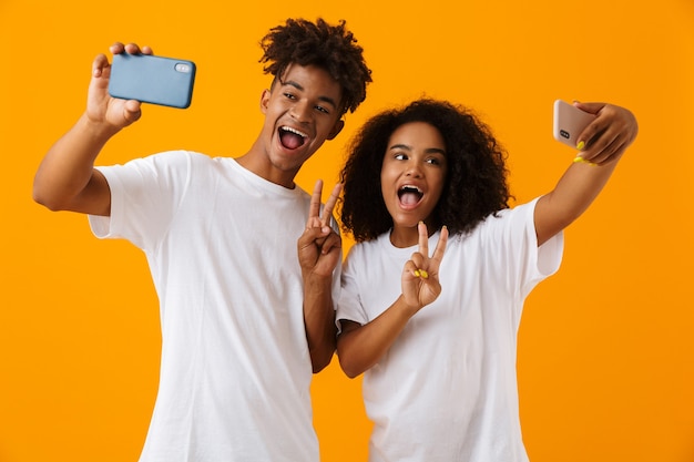 Feliz pareja africana joven linda aislada sobre el espacio amarillo que muestra el gesto de la paz hace selfie por teléfonos móviles.