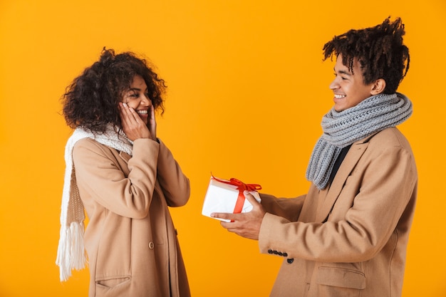 Feliz pareja africana vistiendo ropa de invierno que se encuentran aisladas, dando regalos