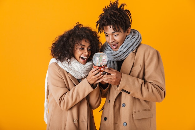 Feliz pareja africana vistiendo ropa de invierno que se encuentran aisladas, sosteniendo snowglobe