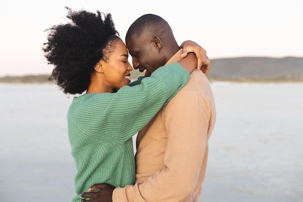 Feliz pareja afroamericana abrazándose y sonriendo en la playa al atardecer. Verano, convivencia, romance y vacaciones, inalterados.