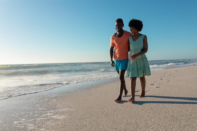 Feliz pareja afroamericana caminando juntos en la playa contra el cielo azul claro en un día soleado