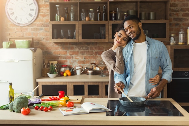 Feliz pareja afroamericana cocinando y probando alimentos saludables en su cocina tipo loft en casa. Mujer y hombre abrazando mientras prepara ensalada de verduras.