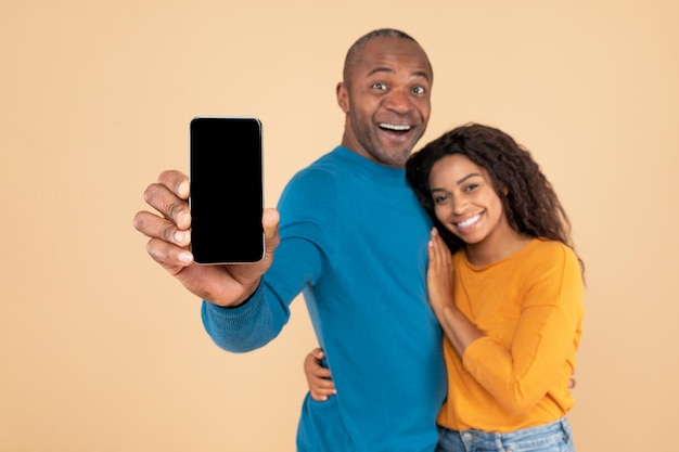 Feliz pareja afroamericana mostrando teléfono celular con pantalla en blanco abrazando y sonriendo a la maqueta de la cámara