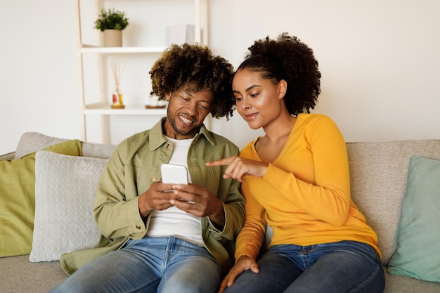 Feliz pareja afroamericana usando un teléfono inteligente señalando con el dedo en casa