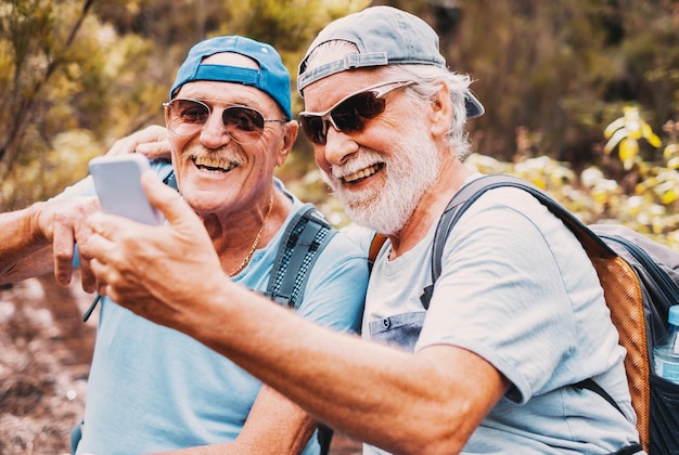 Feliz pareja de amigos mayores con gorras y mochilas en una excursión de montaña mirando el teléfono móvil riéndose mientras se sientan a descansar disfrutando de un estilo de vida saludable en la naturaleza