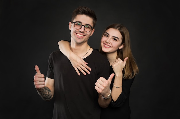 Feliz pareja amor emocionado sonriendo sosteniendo el pulgar hacia arriba gesto, hermosa joven y mujer sonríen, aislado sobre pared negra